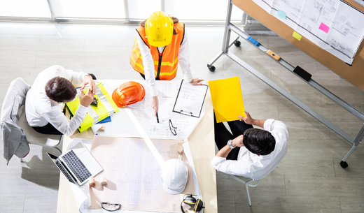 Construction manager in a yellow safety vest leading a project planning and safety meeting with engineers reviewing blueprints and documents.