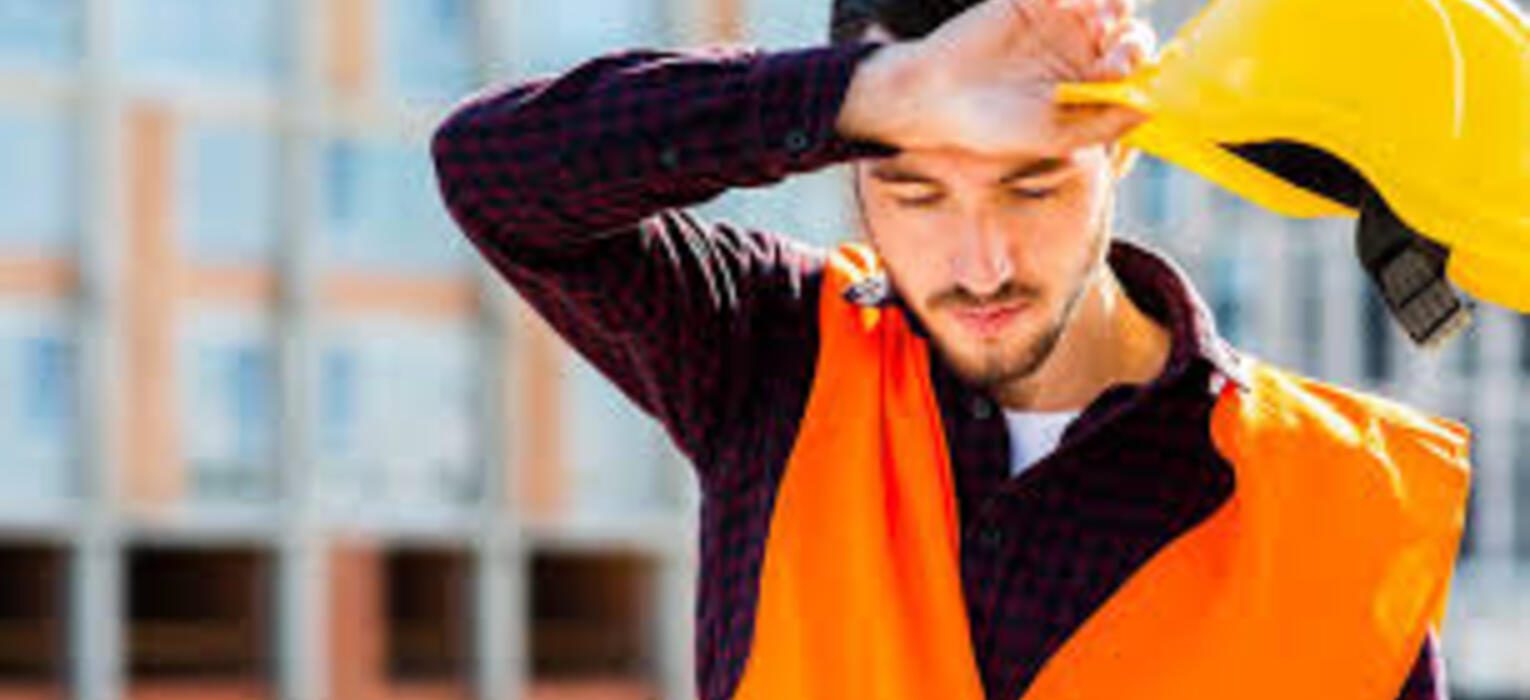 Man in an orange safety vest wiping sweat from his forehead while holding a yellow hard hat, illustrating heat stress awareness in construction settings.