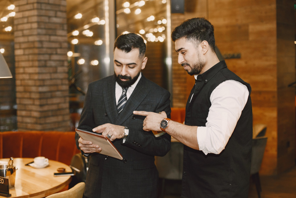 Two hospitality staff members discussing training materials on a tablet in a modern restaurant setting, emphasizing teamwork and communication for effective emergency preparedness and safety training.