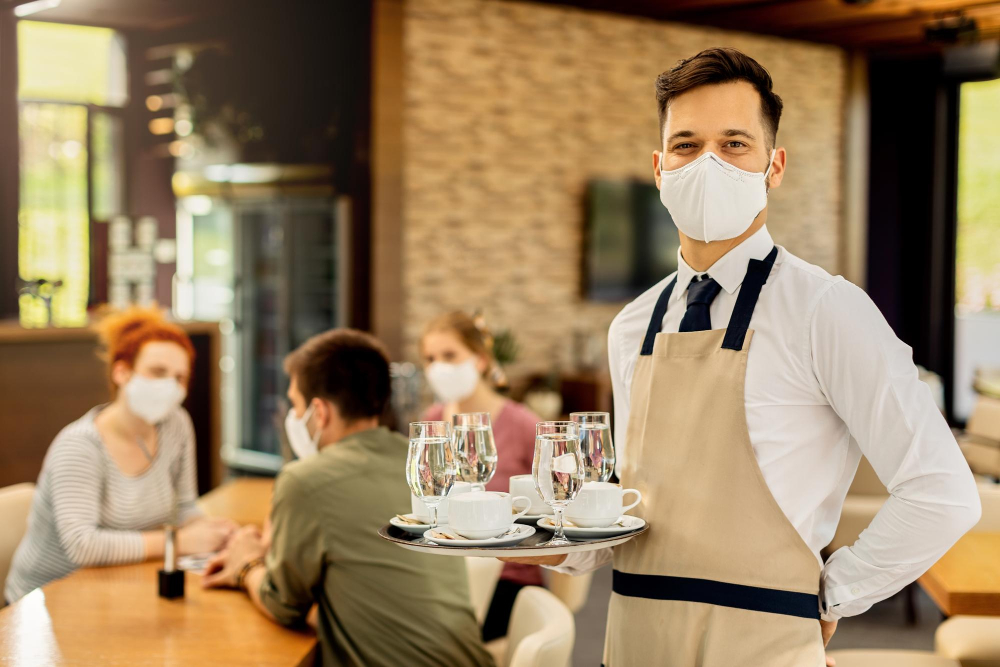 Waiter in face mask serving drinks on a tray to guests in a restaurant setting, emphasizing safety and hospitality training.