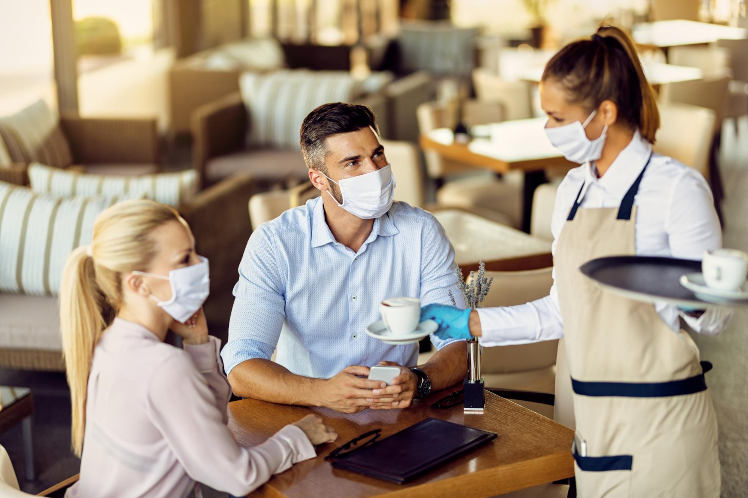 Hospitality staff serving coffee to guests in a restaurant setting, emphasizing safety protocols with masks and gloves, reflecting training in emergency preparedness and guest well-being.