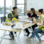 Three individuals in safety vests discussing safety training strategies at a table in a bright, modern workspace, emphasizing workplace safety and emergency preparedness.