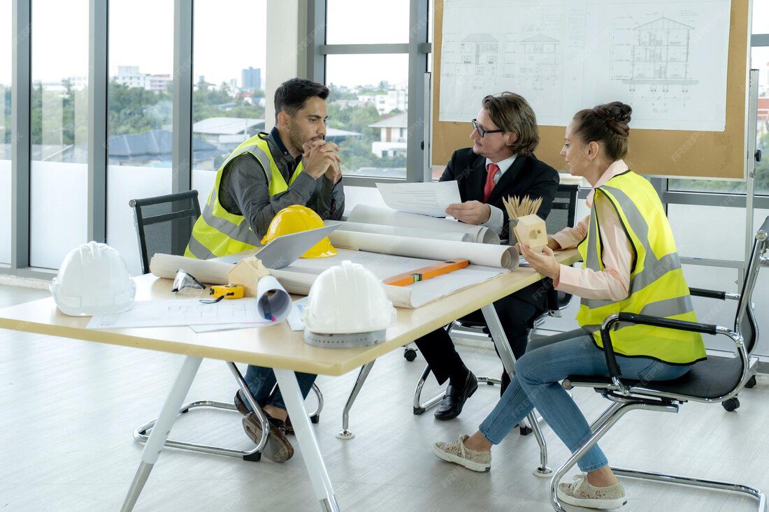 Three professionals in safety vests discussing project plans at a table with construction documents and tools, emphasizing workplace safety training and accident prevention.
